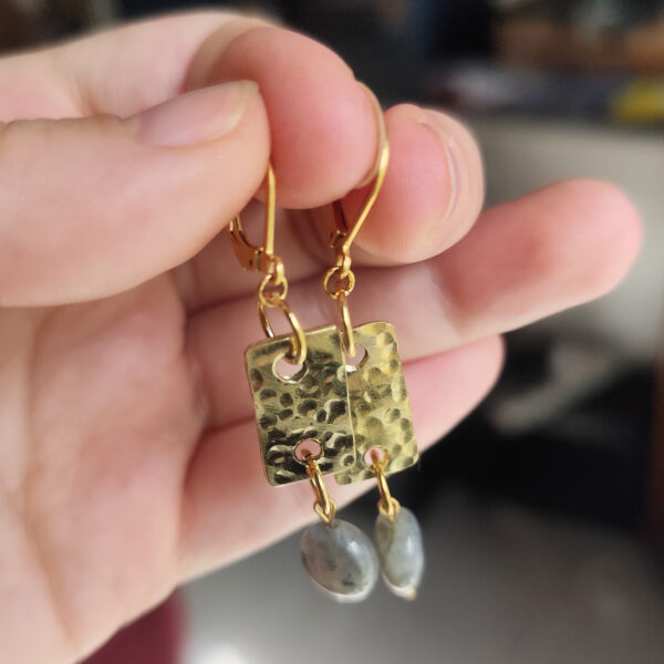 Main qui tient une paire de boucles d'oreilles rectangulaires en laiton martelé et perles de labradorite