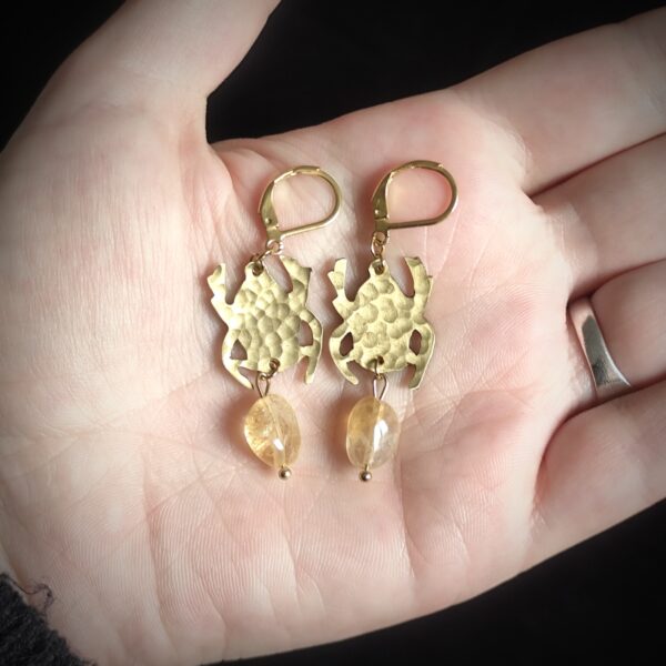 Photo d'une main portant une paire de boucles d'oreilles en forme de scarabée en laiton martelé et perle en citrine
