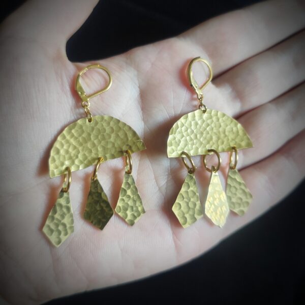 Main portant une paire de boucles d'oreilles avec arc de cercle et pampilles en laiton martelé